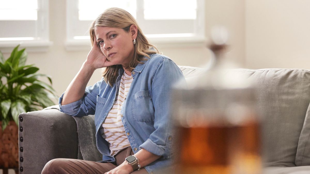 Eine Schwester sitzt auf dem Sofa und starrt quer durch den Raum auf eine Flasche mit Alkohol.