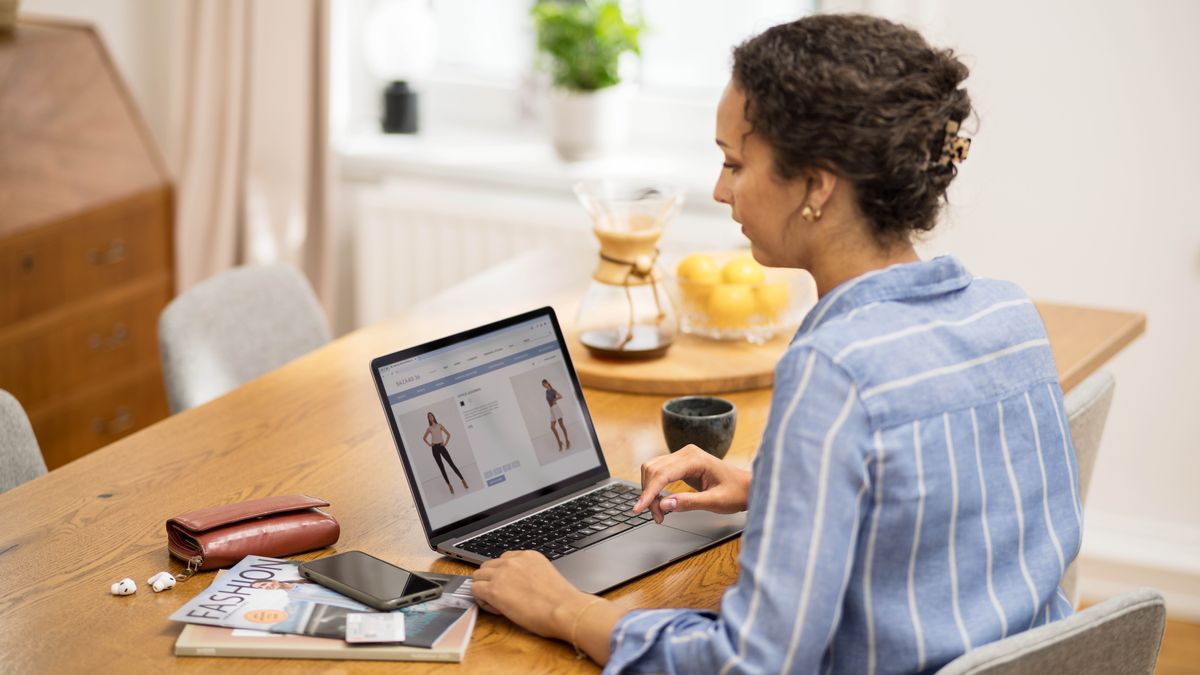 Seorang saudari mencari pakaian yang sopan di toko online. Di sebelah laptopnya, ada majalah pakaian.
