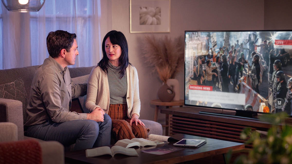 A couple watching a TV news report of civil unrest. They have a Bible and other publications on a table in front of them.