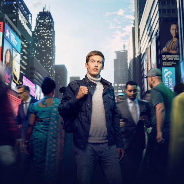 A young man standing on a busy city street. People of different nationalities and backgrounds are passing by him. He is looking up as if seeking direction.