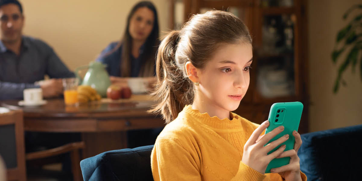 女の子がスマホに夢中になっている。両親は女の子が朝食の席に着くのを待っている。
