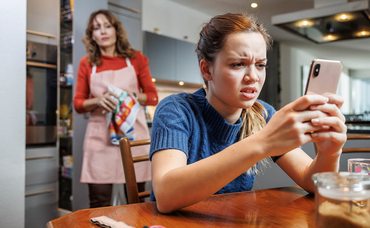 10代の女の子がスマホを見ていらいらしている。その様子を母親が離れて見ている。