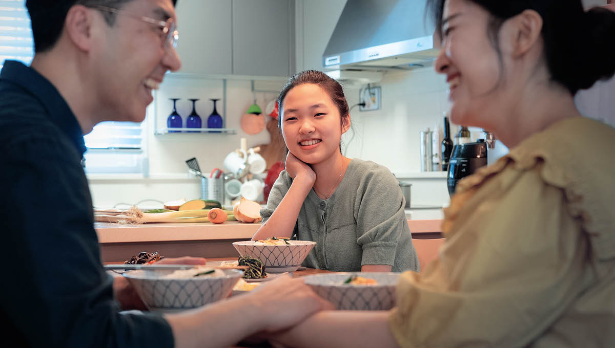 夕食のテーブルで10代の娘がうれしそうに両親を見ている。父親が母親の手を握り，2人で笑っている。
