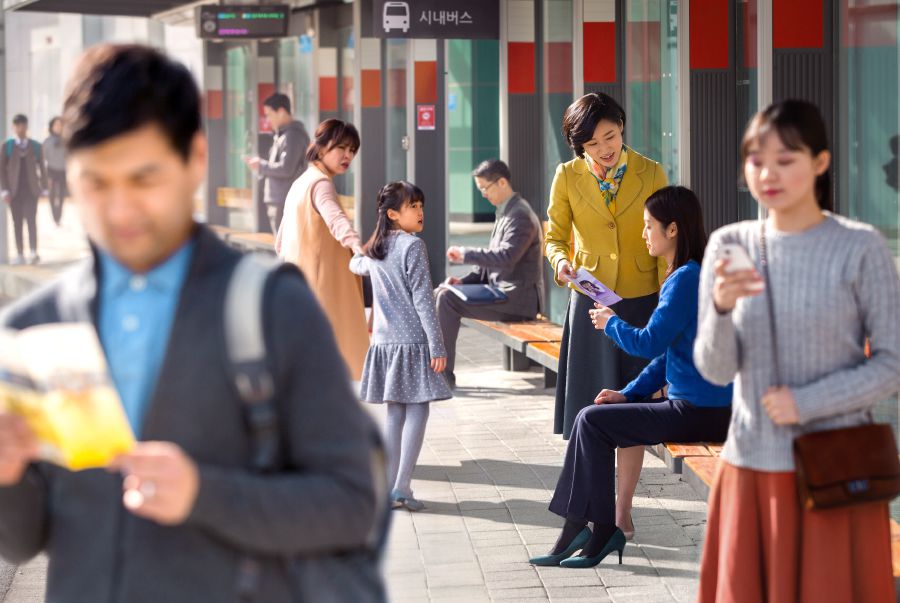 1人の姉妹がバス停で女性にパンフレットを渡している。周囲のほとんどの人は無関心に見える。