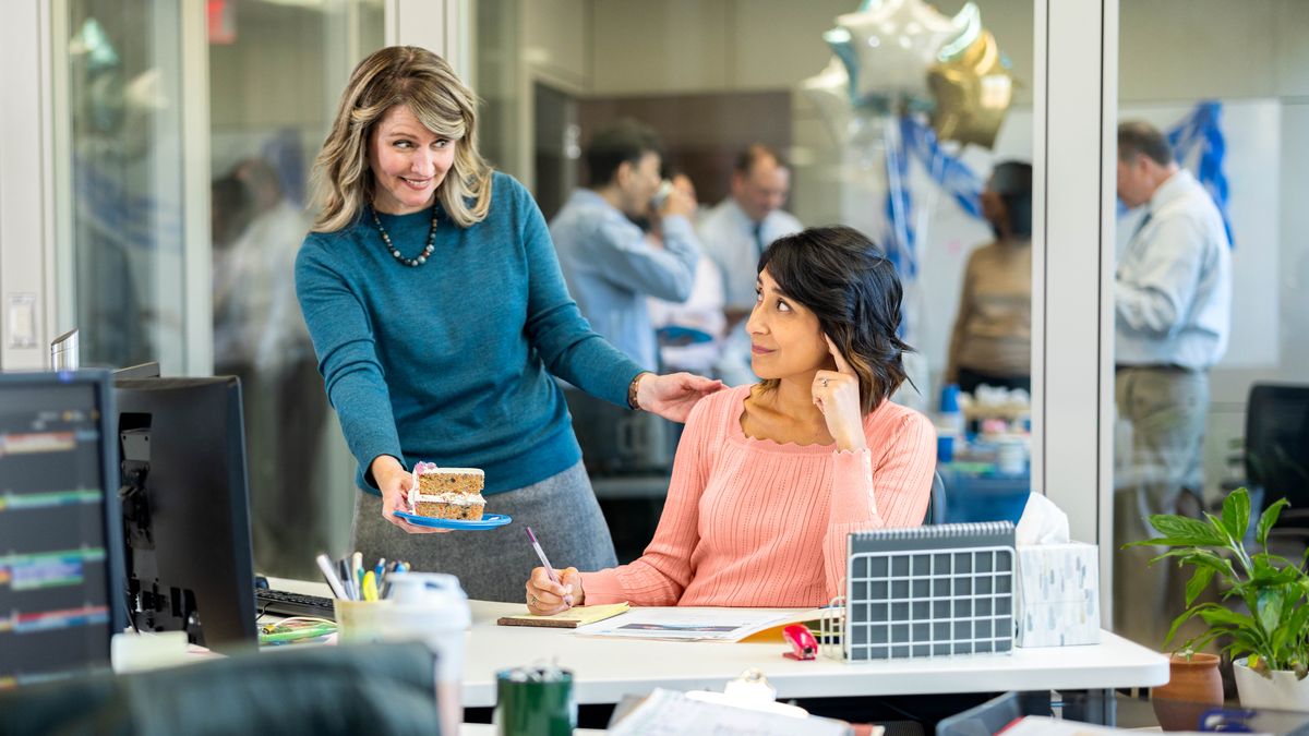 The sister workmate giving her birthday cake to her desk. Other people eating, drinking, and celebrating in the other office.