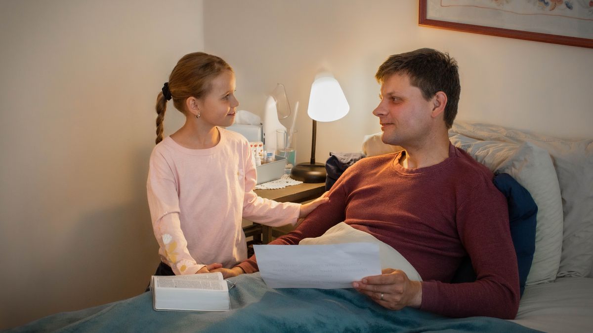One brother laying down on the bed holding one sheet of paper and his small daughter holding his hand.