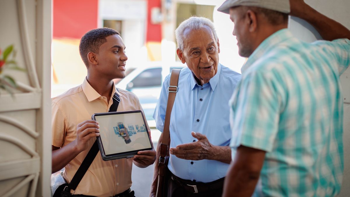 The young brother and the older brother preaching from house-to-house. The older brother talking with the man while the young brother holding the tablet that playing the video.
