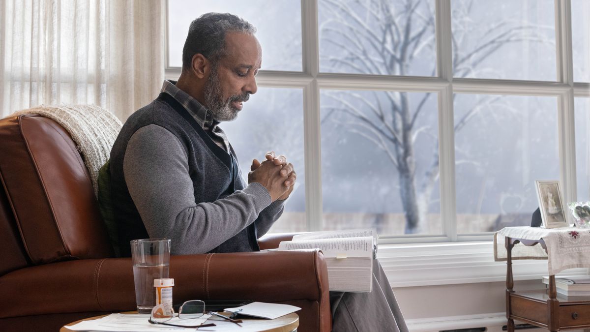 The old brother sitting down in his house praying. The Bible open on his lap, and his one medicine bottle on the table near him.