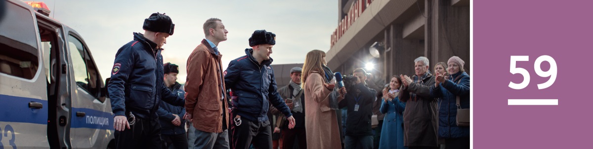 59 pamoka. Antrankiais surakintą Jehovos liudytoją veda du policijos pareigūnai; juos stebi žurnalistai ir bendratikiai.