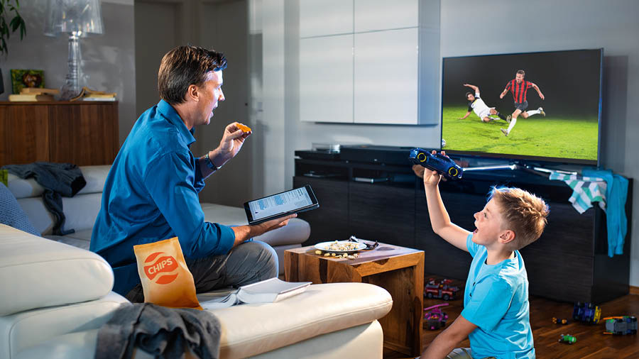 Un hermano comiendo y viendo la televisión mientras hace su estudio personal. Su hijo está a su lado jugando.