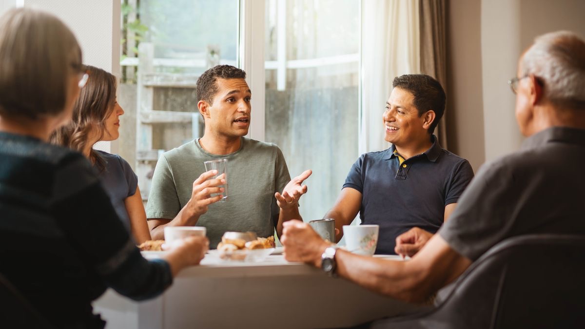 Alguns irmãos e irmãs tomando uma refeição juntos. Uma irmão está falando enquanto outros prestam atenção no que ele diz.