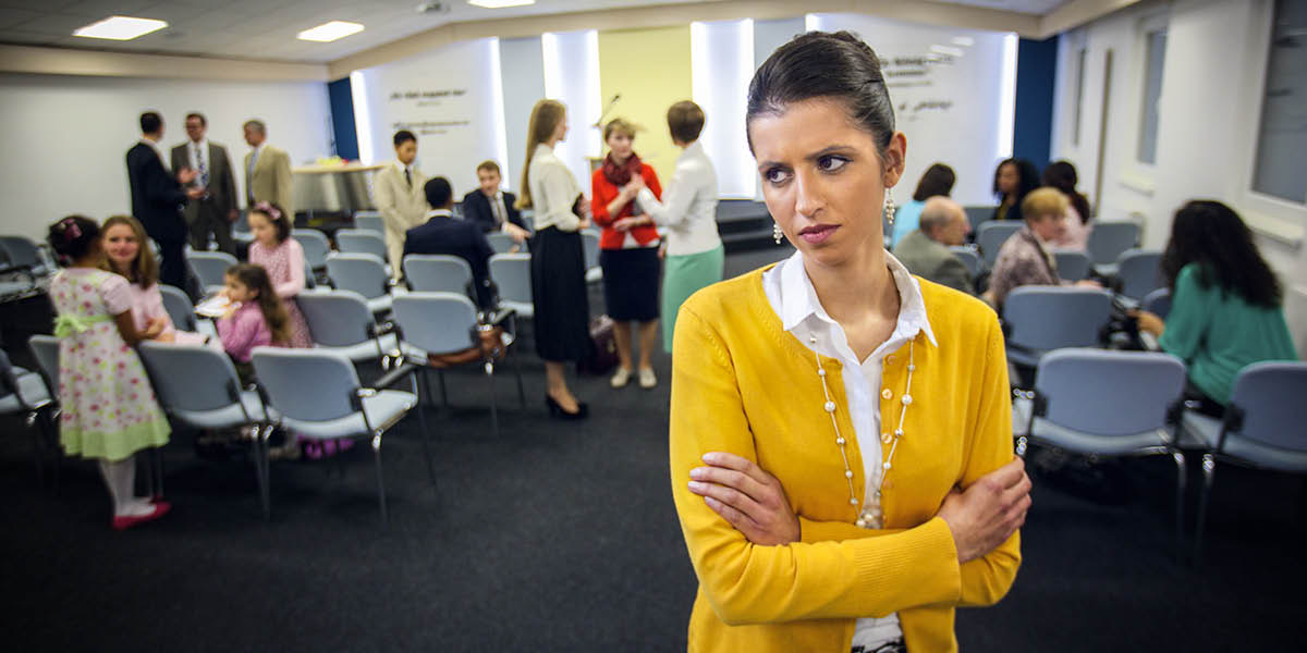Een ongelukkige zuster staat met haar armen over elkaar op afstand van de aanwezigen in een Koninkrijkszaal.