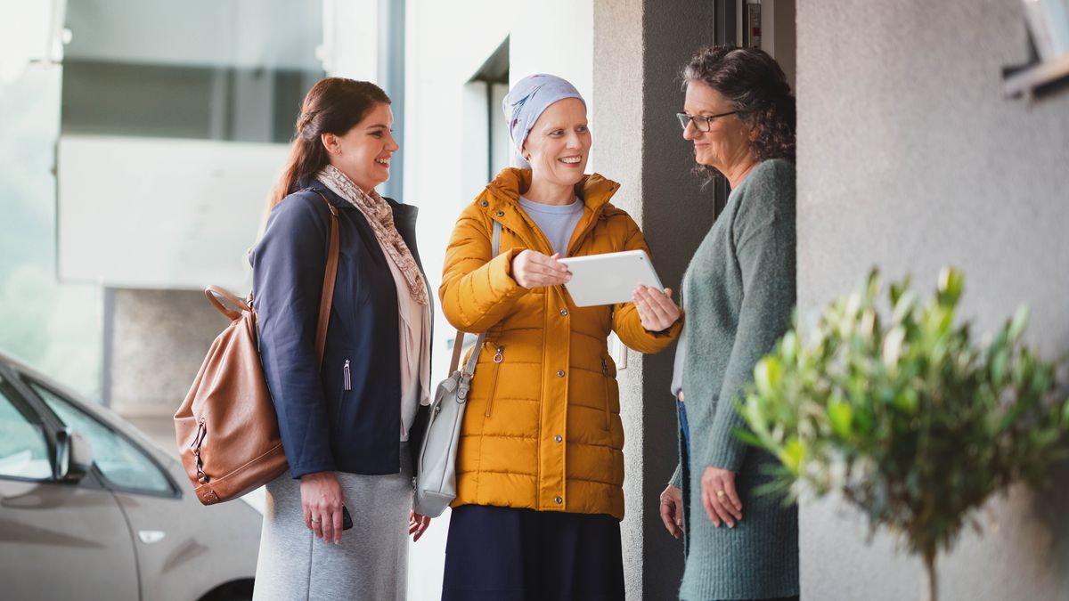 Een zuster die een ernstige ziekte heeft, is met een vriendin in de velddienst. Ze kijkt blij terwijl ze een vrouw aan de deur iets op haar tablet laat zien.