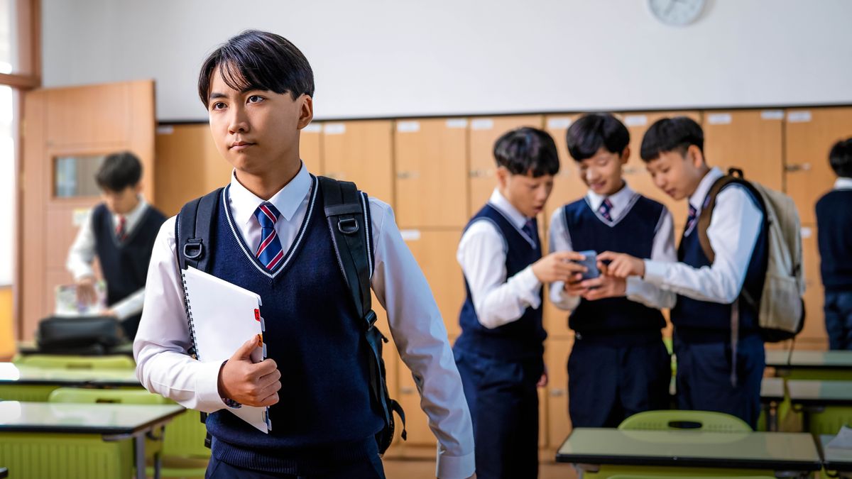 Een jonge broeder loopt weg bij zijn klasgenoten, die zich vergapen aan iets op een smartphone.