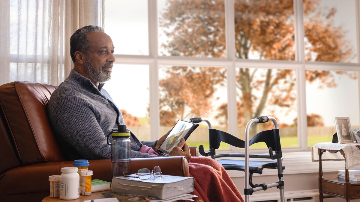 De oudere broeder zit in de herfst thuis na te denken over de afbeelding van het paradijs op zijn tablet. Naast hem staat een looprek en er staan meerdere potjes medicijnen.