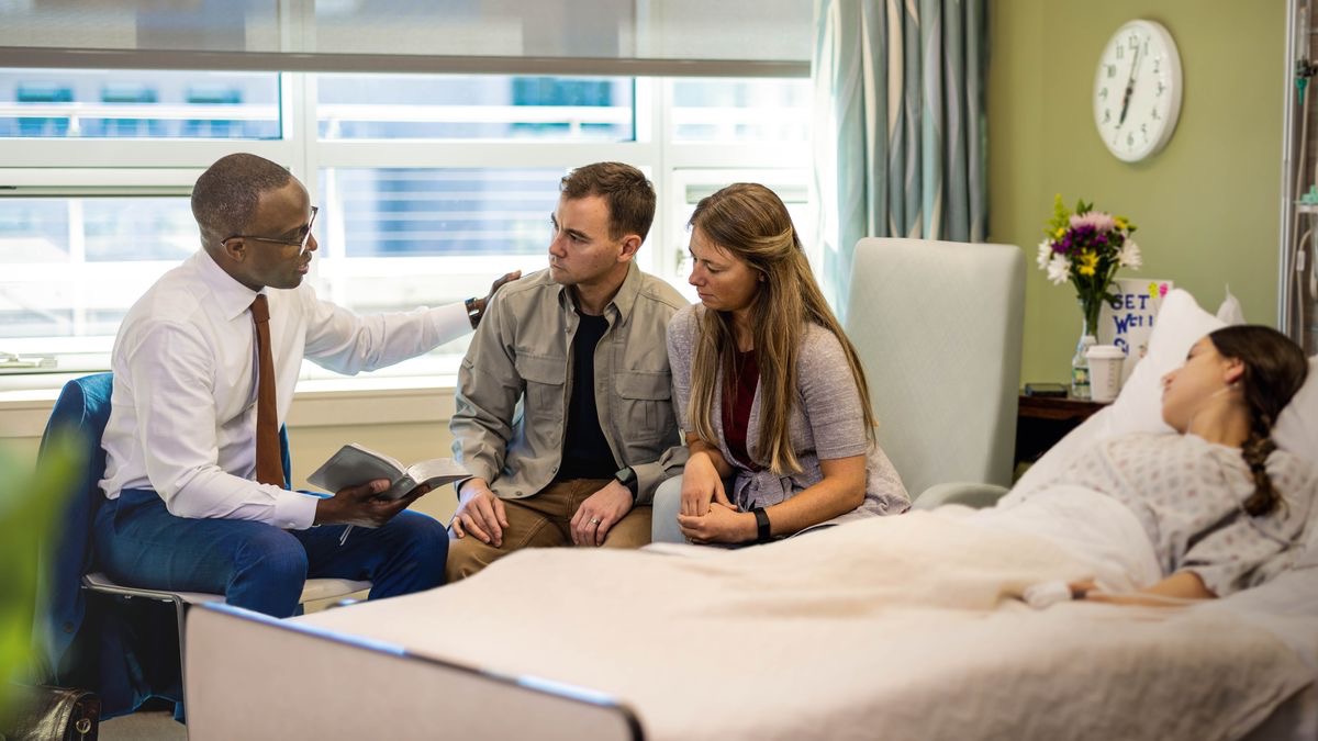 Un ansiano ta usa Beibel pa konsolá un pareha. Un hobensita ta drumi riba kama di hospital.