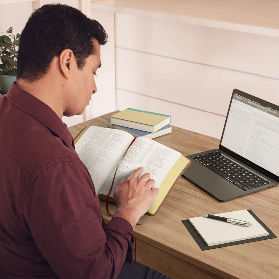 Man hia iusim Bible and laptop for duim research. Wanfala notepad and olketa buk lo desk blo hem.