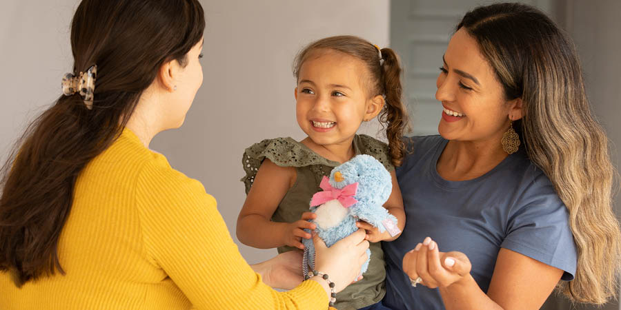 Mama trzyma na rękach swoją małą córeczkę, która dostaje w prezencie maskotkę. Obie uśmiechają się z wdzięcznością.