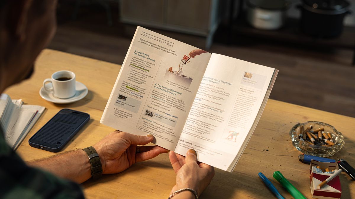Mężczyzna czyta 40 lekcję książki „Już zawsze ciesz się życiem!”. Na jego biurku leży paczka papierosów i popielniczka pełna niedopałków.