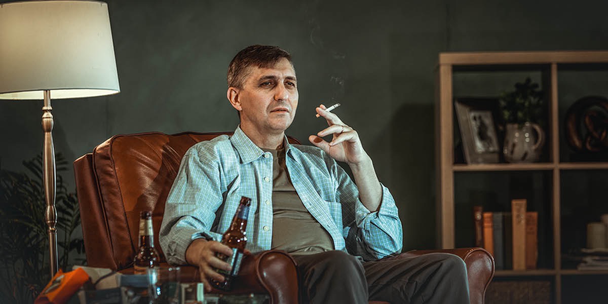 Mężczyzna siedzi w pokoju z papierosem w jednej ręce i butelką piwa w drugiej ręce.