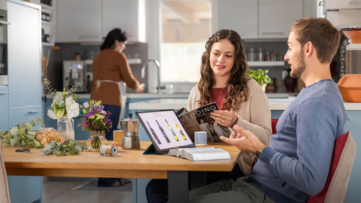 Um casal de noivos a falar sobre se devem servir álcool ou não na sua festa de casamento. Eles têm uma Bíblia aberta colocada sobre a mesa, juntamente com um tablet que está a mostrar imagens de vários tipos de vinho.