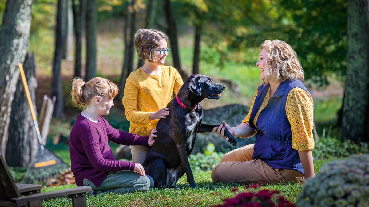 Uma mãe e as suas duas filhas conversam num parque e fazem festas a um cão.