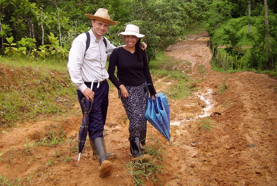 Donald e Nuria numa estrada lamacenta, com chapéus, mochilas, guarda-chuvas e botas de borracha.