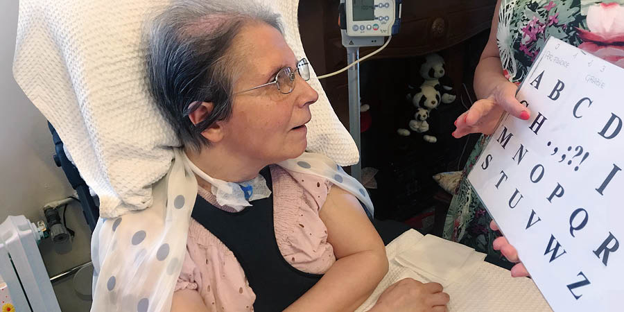 Virginia a olhar para o quadro com o alfabeto, enquanto a outra pessoa aponta para uma das letras.