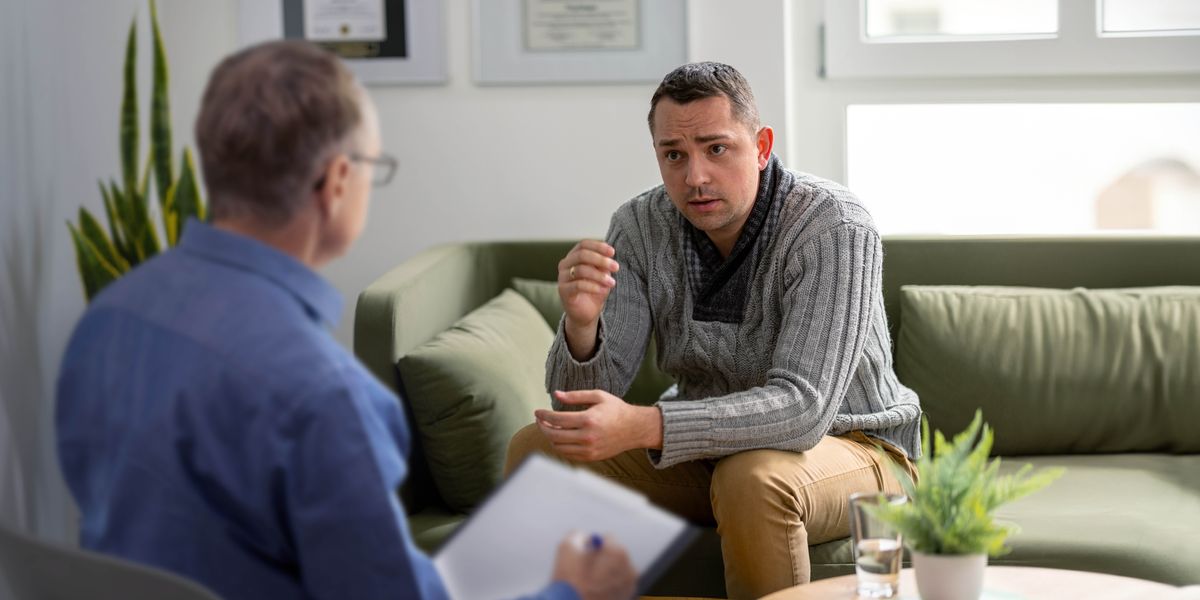 O marido está sentado no sofá e fala com um profissional de saúde.