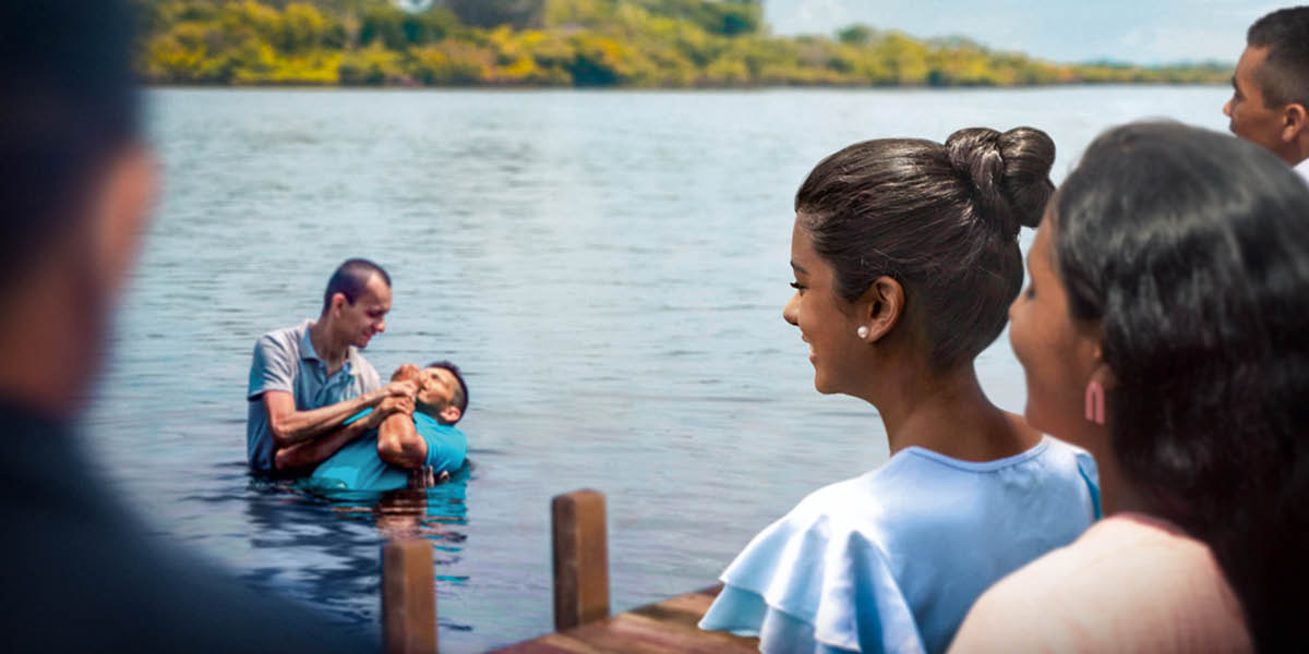 Um homem a ser batizado num lago, enquanto outros assistem a partir da doca.