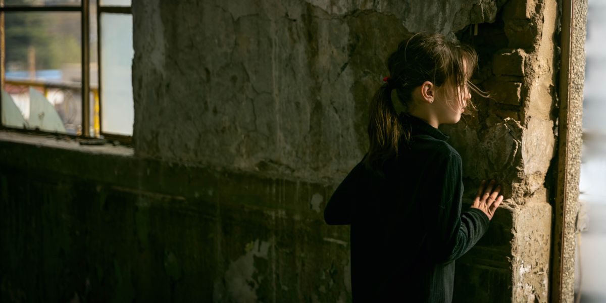 Uma menina a espreitar atrás de uma parede de um edifício destruído.