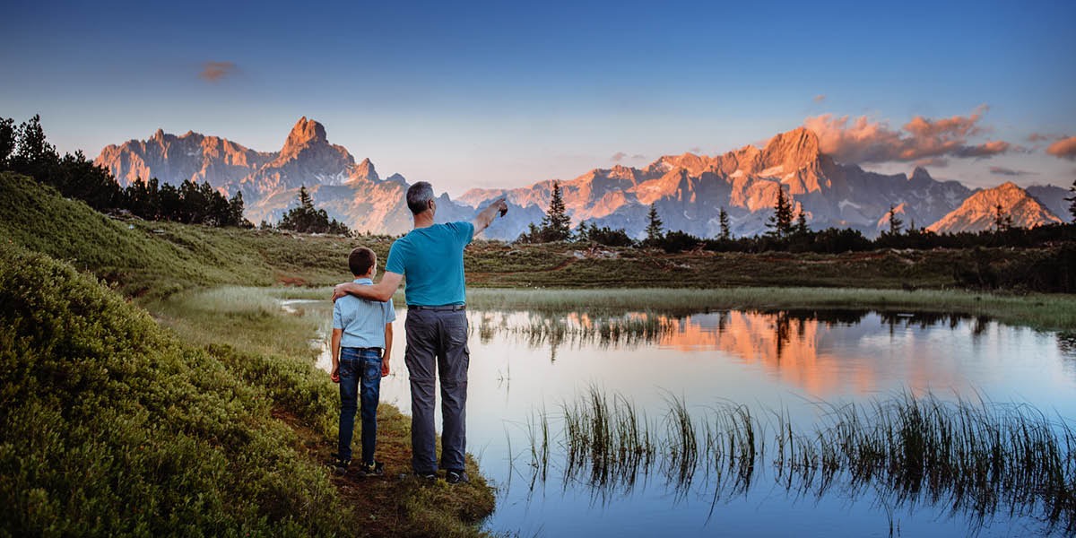 Um pai e um filho ao pé de um lago a olharem para as montanhas.