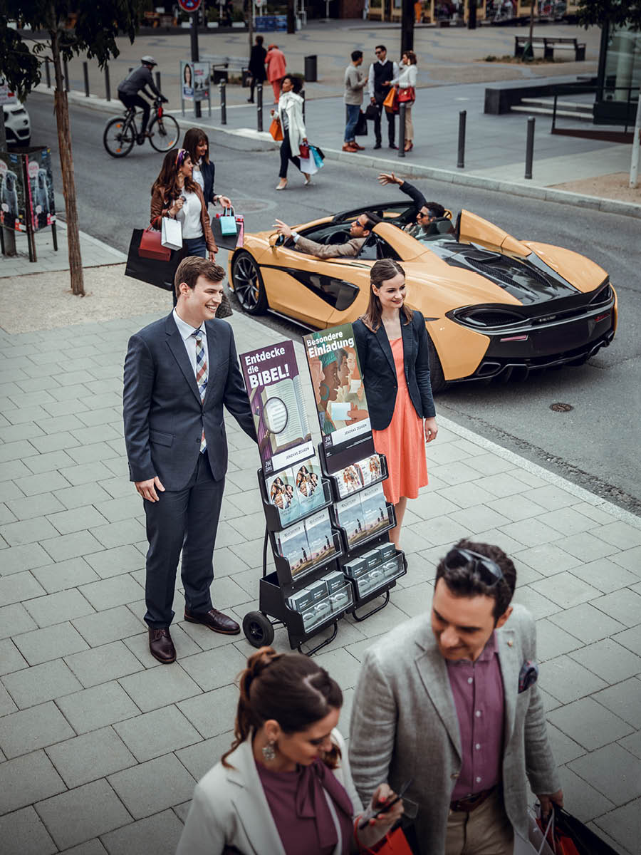 Um casal participa alegremente no testemunho público, enquanto as pessoas à sua volta andam às compras e conduzem carros caros.