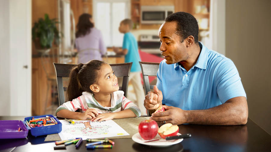 Um pai e uma filha conversam. Ela está a colorir um desenho de uma macieira e o pai está a apontar para algumas maçãs que estão na mesa.