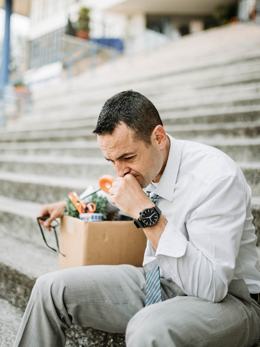 Um homem está triste porque foi demitido do trabalho. Ele está sentado do lado de fora da empresa e está com uma caixa com os seus itens pessoais.