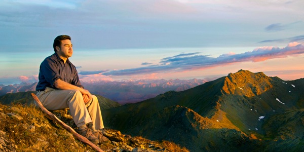 Um homem sentado no topo de uma montanha olha a paisagem em volta e aprecia um lindo céu
