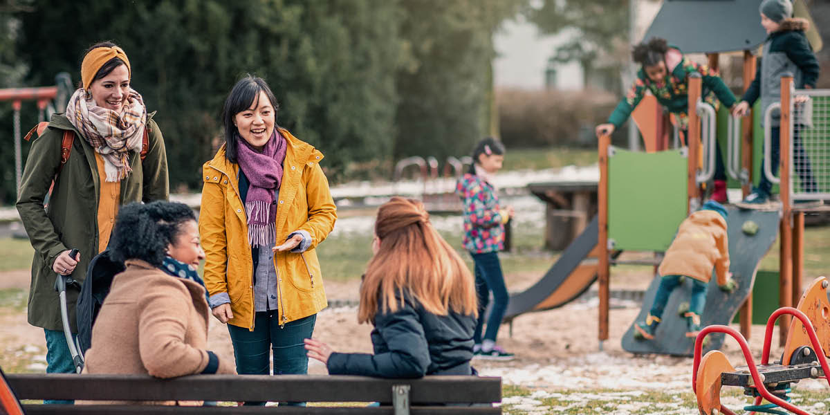 Quatro mulheres de raças diferentes conversando e rindo enquanto os filhos delas brincam num parque.