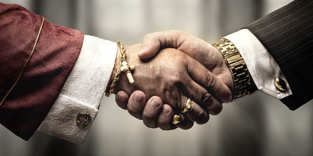 Um líder religioso apertando a mão de um homem de negócios. Os dois estão usando joias de ouro.