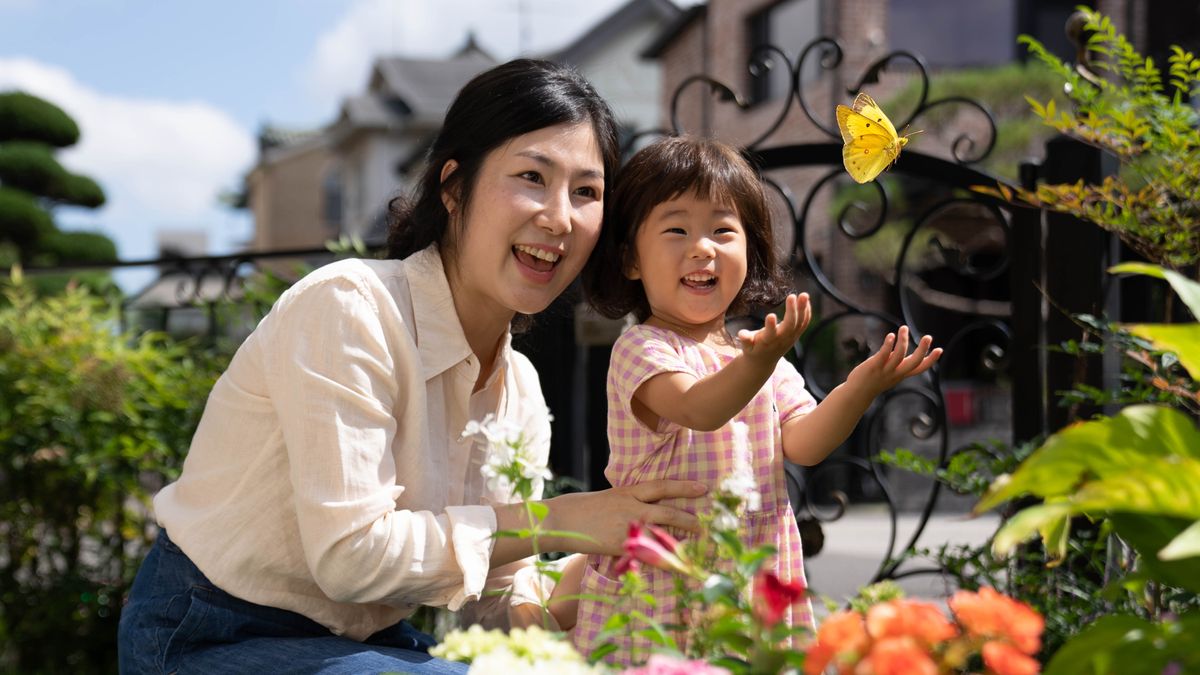 Uma mãe com sua filhinha olham felizes para uma borboleta no jardim de casa.