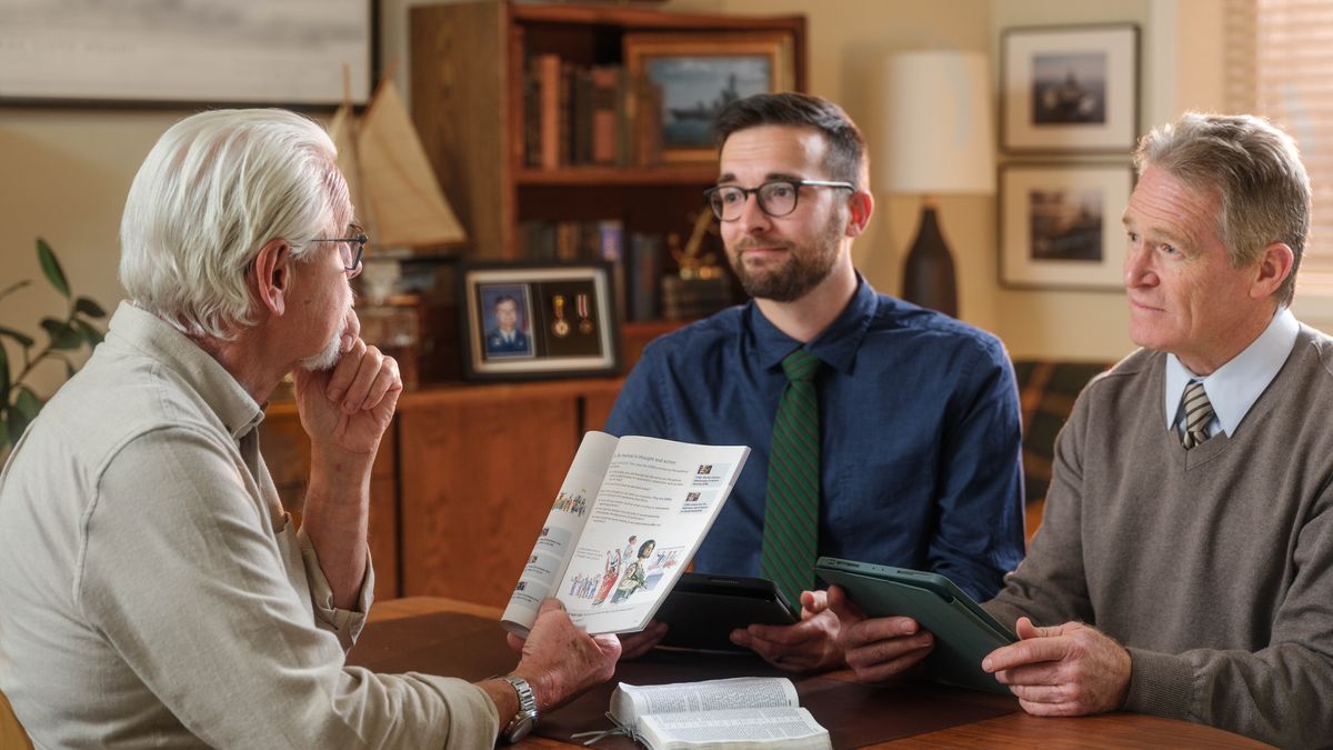 O homem idoso da imagem anterior estudando o livro “Seja Feliz para Sempre!” com dois irmãos. Perto deles, há um quadro com medalhas e condecorações militares.