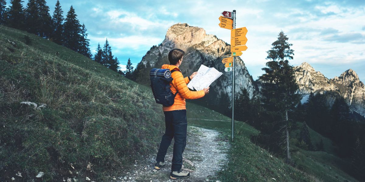 O mesmo homem dos outros artigos está parado na trilha de uma montanha olhando para um mapa. Na frente dele, tem um poste com várias placas de direção. Ao fundo, dá para ver o topo de uma montanha.