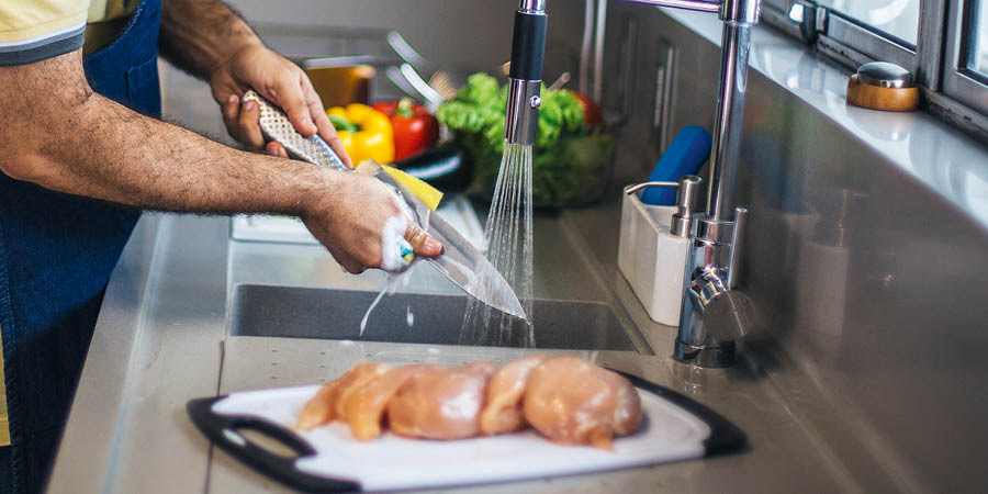 Un hombre lavando un cuchillo con agua y jabón. El pollo crudo y las verduras están en dos tablas diferentes.