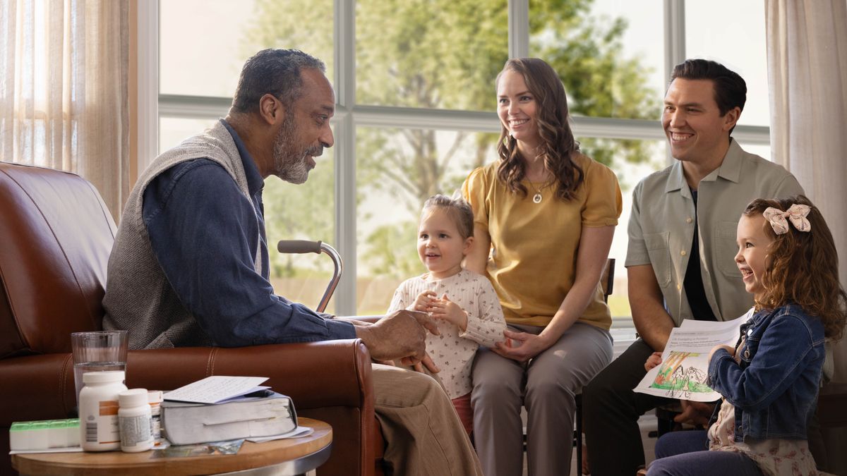 El hermano mayor de la imagen anterior sentado en su casa durante la primavera. Conversa sonriendo con dos niñas pequeñas y sus padres. Junto a su bastón unas pocas botellas de medicamentos. Una de las niñas le muestra un dibujo que hizo del Paraíso.