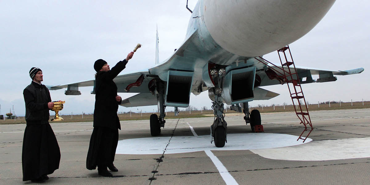 A priest blessing a military fighter jet, sprinkling holy water on it.