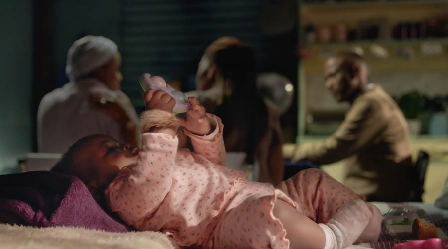 Um bebê numa cama com um amuleto no pulso. A mãe do bebê está conversando com o casal que estuda a Bíblia com ela.