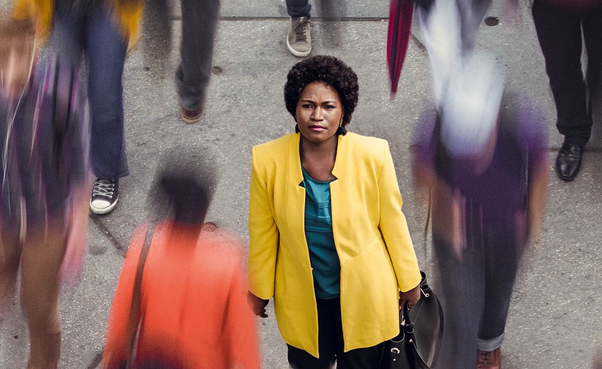 Uma mulher de pé numa rua com várias pessoas passando ao seu redor. Ela olha para cima como se estivesse se perguntando: ‘Será que Deus me vê?’