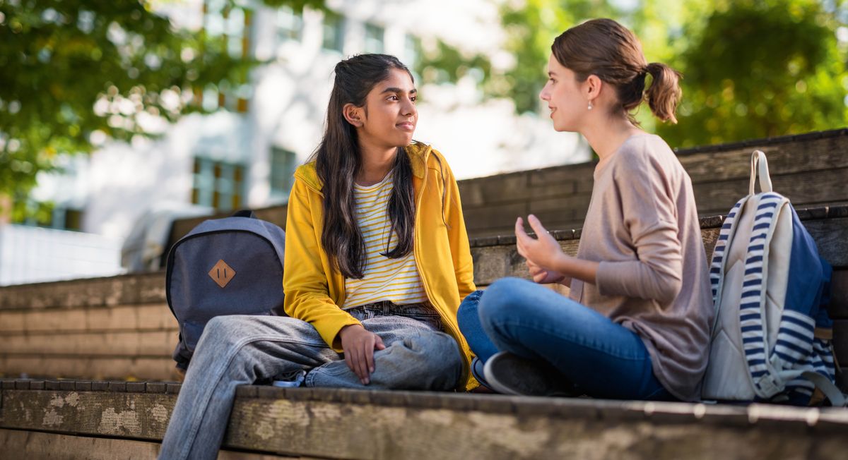 Dy adoleshente janë ulur në një stol në natyrë dhe po shohin njëra-tjetrën. Një nga vajzat po dëgjon me vëmendje ndërsa tjetra shprehet.