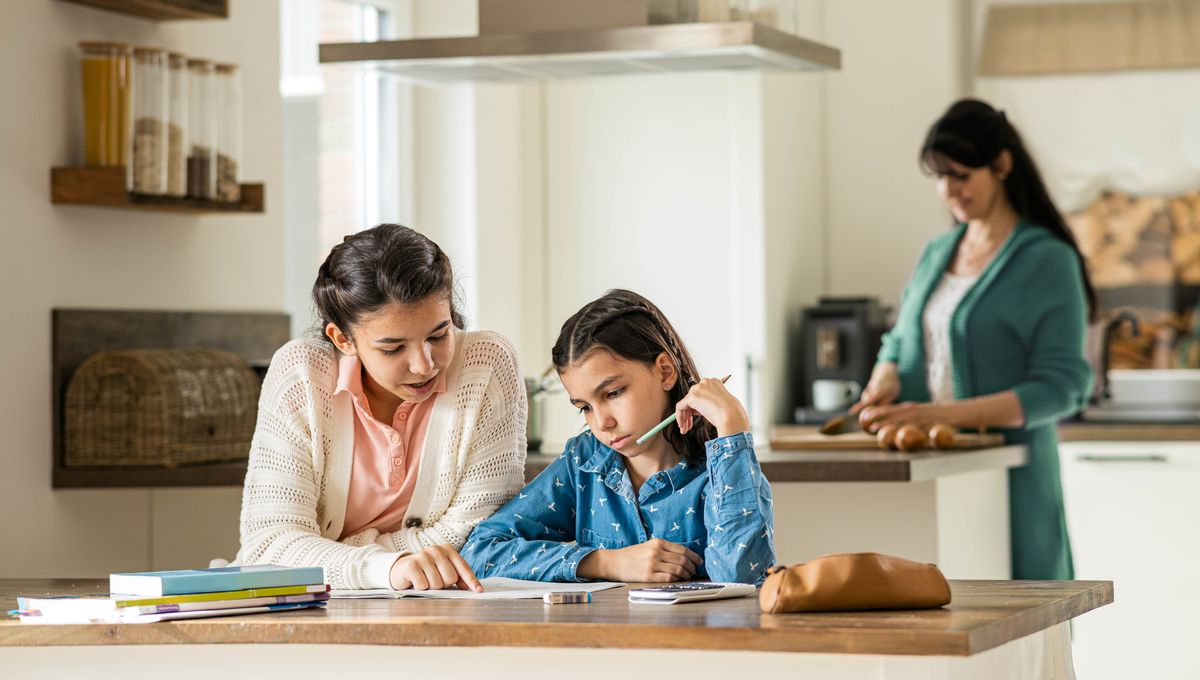 Një adoleshente po ndihmon motrën e saj të vogël me detyrat e shtëpisë, ndërsa mamaja po gatuan në kuzhinë.