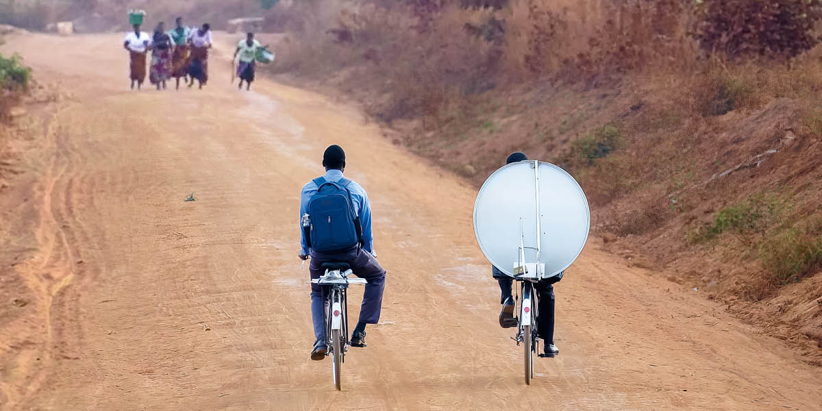 Ndugu wakiendesha baiskeli katika barabara isiyo na lami, wakisafirisha vya satelaiti hadi kwenye makutaniko yao.