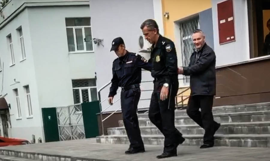 «Дмитрий Михайлов бурадиримиз билән сөһбәт» дегән видеодин елинған бир көрүнүшдегән видеодин елинған бир көрүнүш. Дмитрийниң қолиға кишән селип, уни икки сақчи елип кетиватиду.